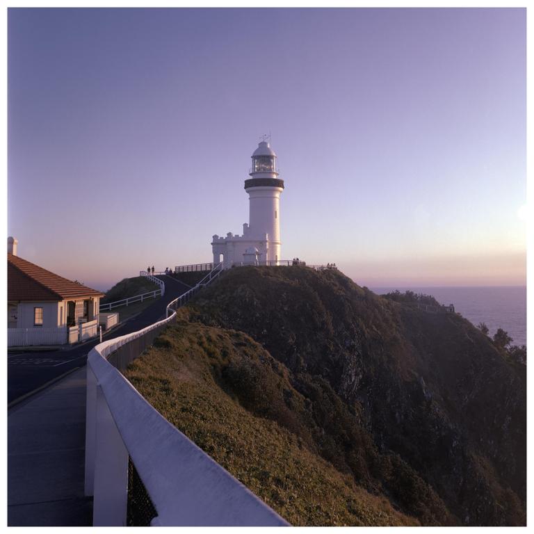 A white lighthouse stands on a grassy hill overlooking the ocean at dusk, with a road and building in the foreground.