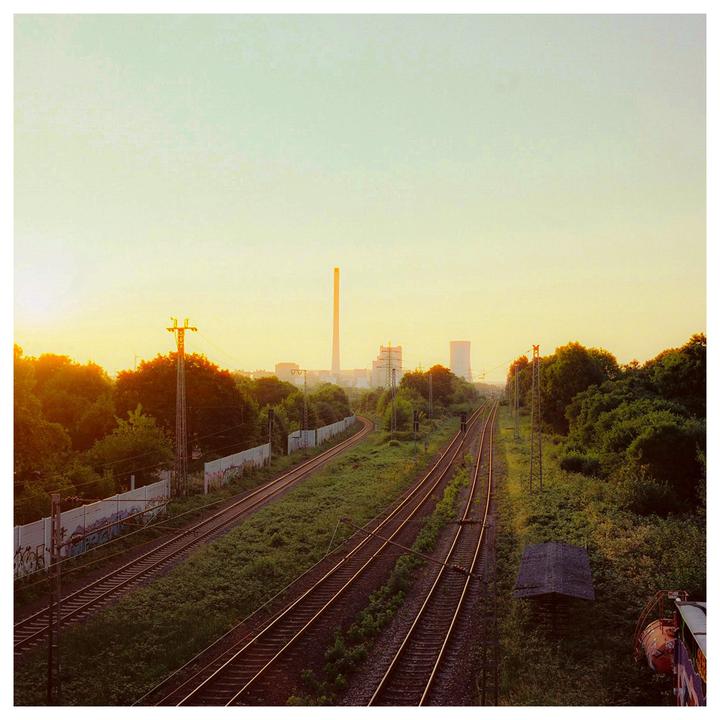 A power plant with a tall smokestack is visible in the distance beyond multiple railroad tracks and green foliage.