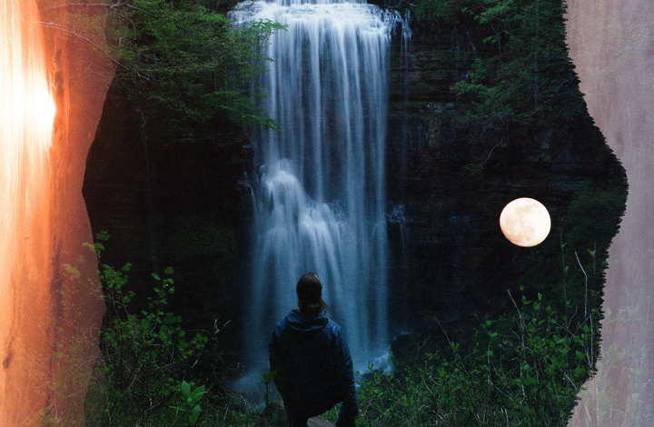 A person stands with their back to the camera, looking at a waterfall under a full moon.