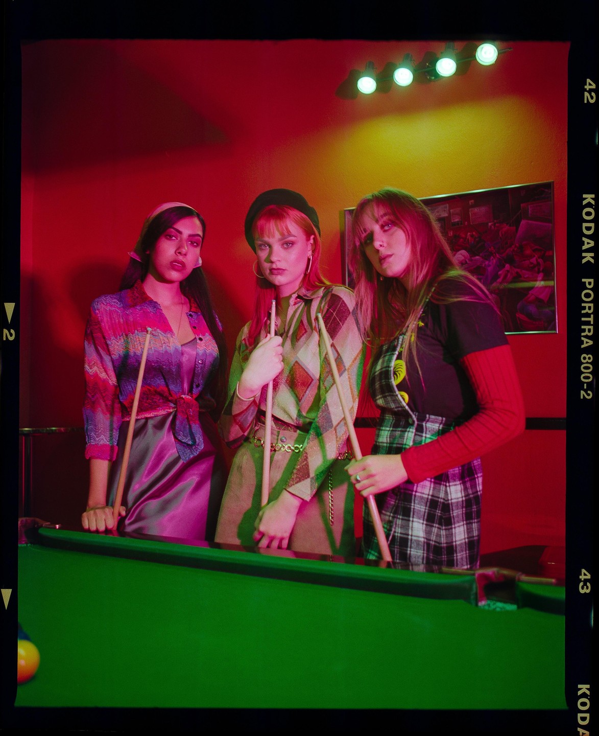 Three women stand around a pool table, holding pool cues, under red and green lighting.