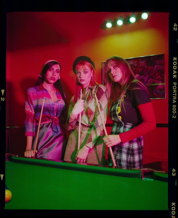 Three women stand around a pool table, holding pool cues, under red and green lighting.