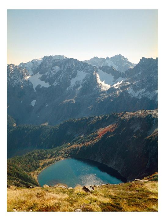 A high-angle landscape shows a blue lake nestled in a valley with snow-capped mountains in the distance.