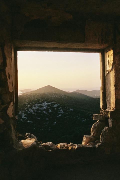 A view of green hills and distant water is seen through a stone window frame at sunset.