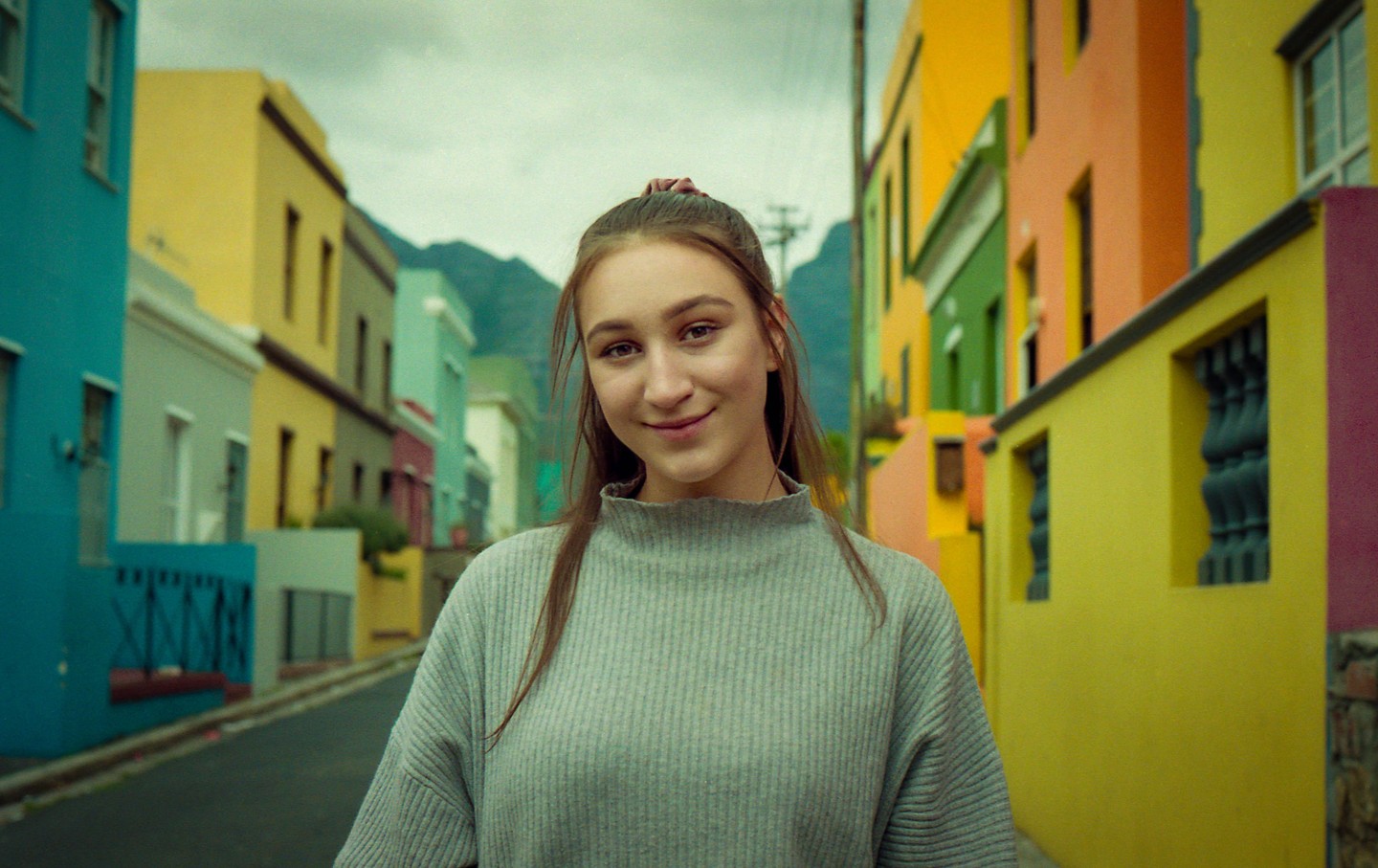 A woman stands on a street lined with colorful buildings, with mountains in the background.