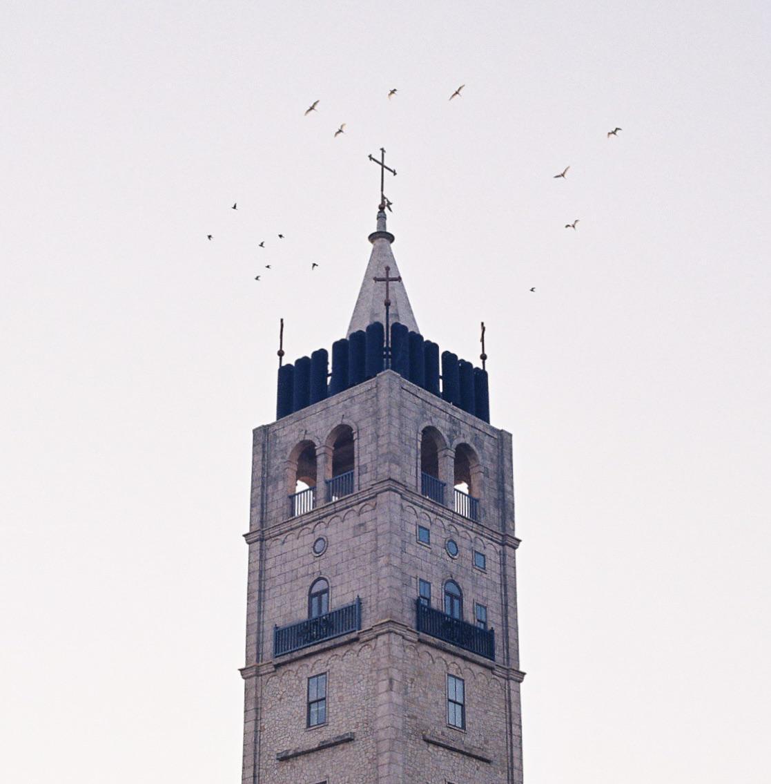 A stone church tower with a cross on top stands against a bright sky with birds flying around it.