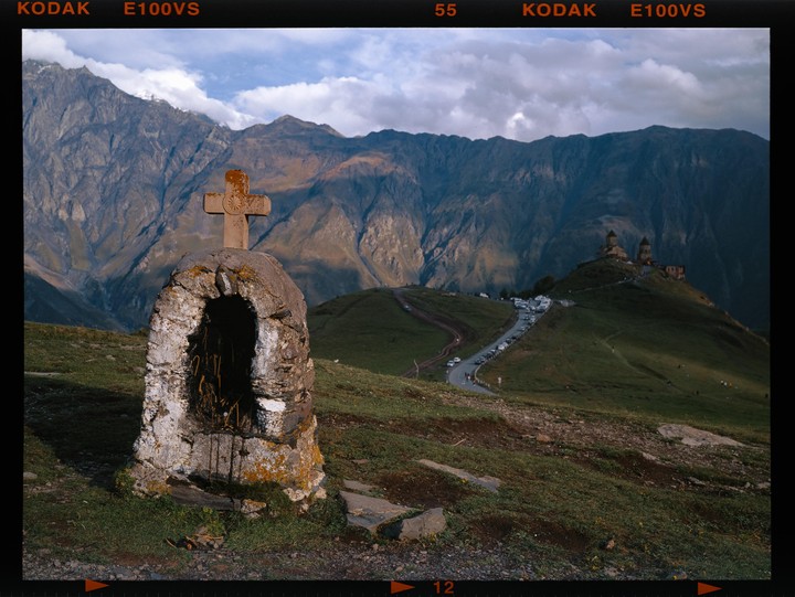 A stone cross and shrine stand in a grassy mountain landscape with a church and road visible in the distance.