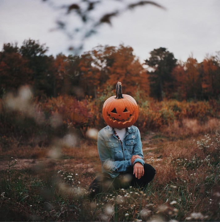 A person wearing a jack-o'-lantern on their head kneels in a field with autumn trees in the background.
