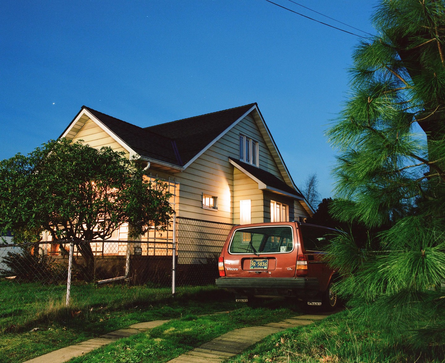 A red Volvo station wagon is parked in a driveway next to a house with illuminated windows at dusk.