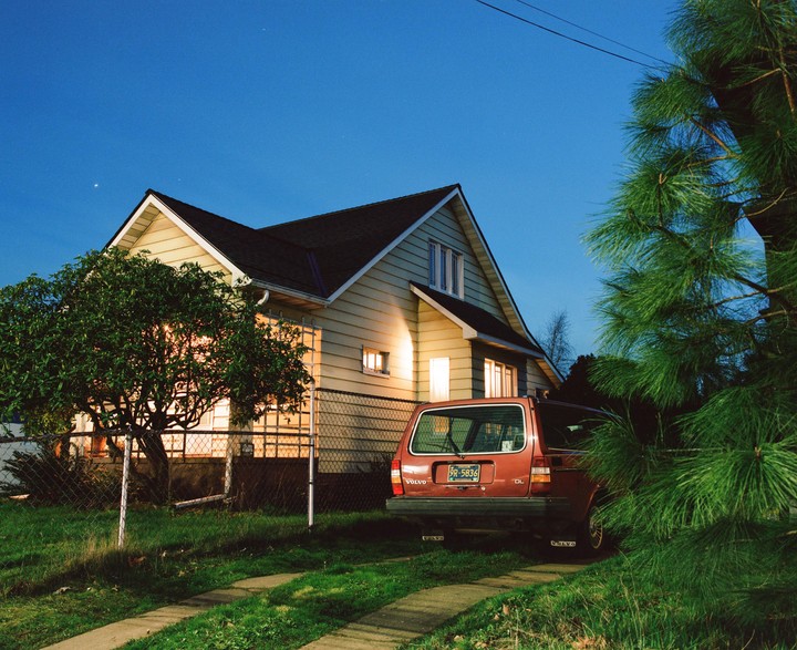 A red Volvo station wagon is parked in a driveway next to a house with illuminated windows at dusk.