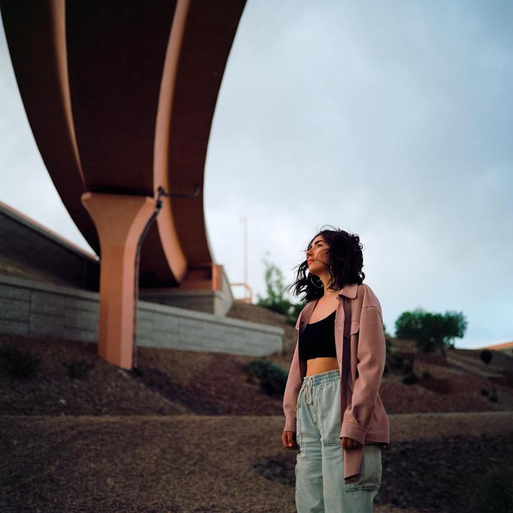 A woman stands under a freeway overpass, looking up at the sky.