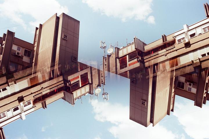 A double exposure shows two concrete buildings against a blue sky with clouds.