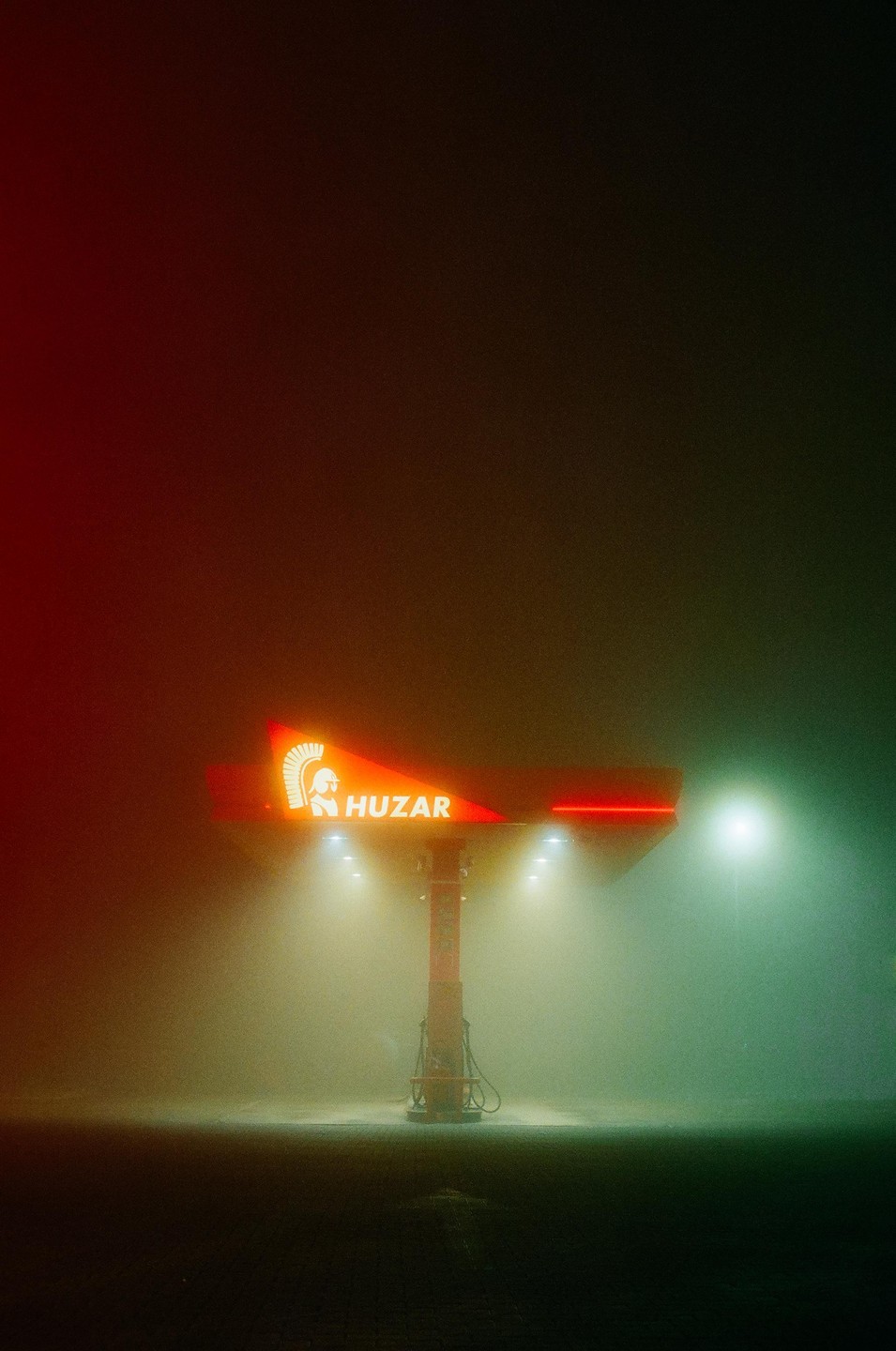 A gas station with a red sign that says "Huzar" is illuminated by bright lights in a foggy night.