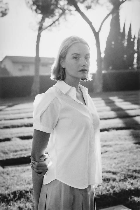 A woman with short hair stands in a grassy area with trees and a building in the background.