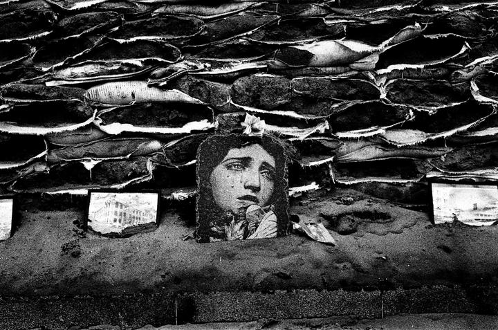 A cutout of a crying woman's face is placed against a wall of sandbags, with a small flower on top of her head.