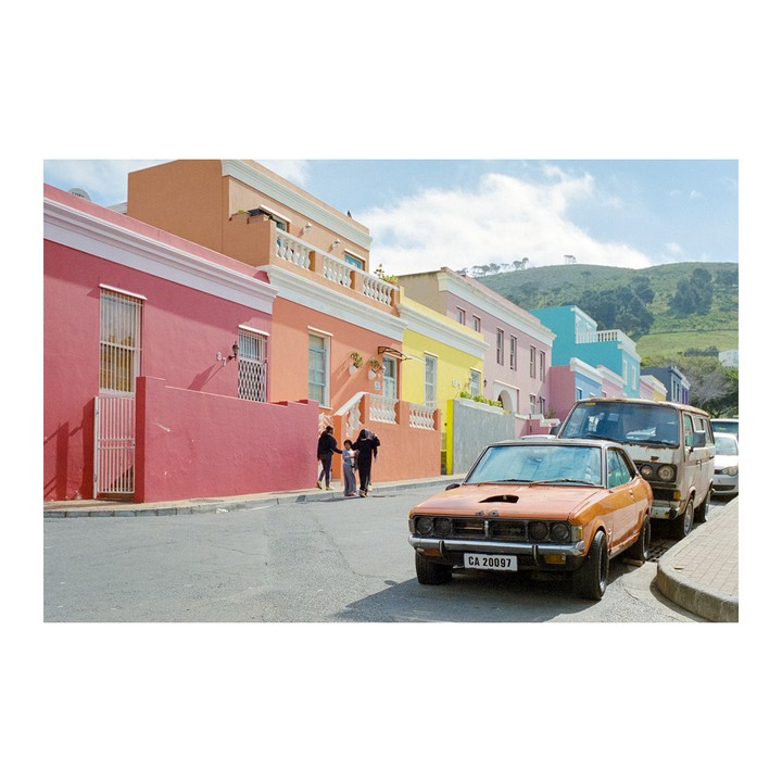 An orange car is parked on a street lined with colorful houses, with people walking on the sidewalk and a green hill in the background.