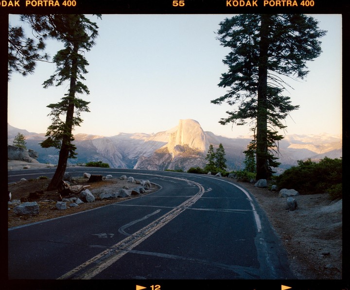 A winding road leads to a view of Half Dome in Yosemite National Park, framed by two tall pine trees.
