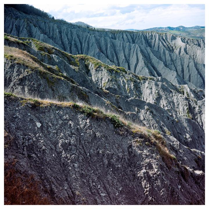 A landscape of badlands with steep, eroded hills and sparse vegetation under a cloudy sky.