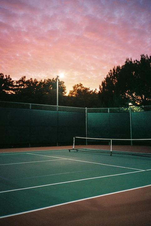 A tennis court is illuminated by artificial lights under a vibrant pink and orange sunset sky.