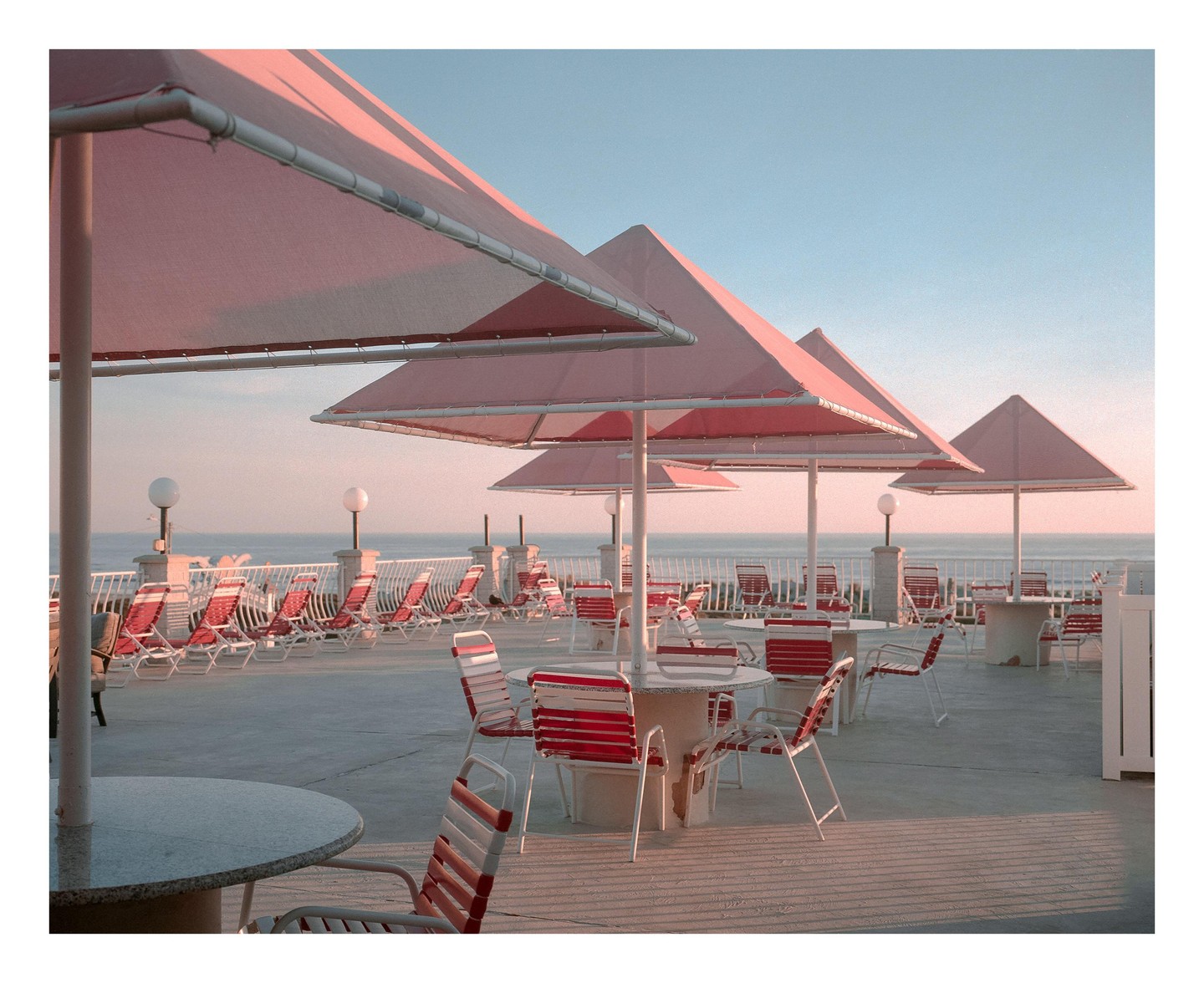 A row of pink square umbrellas shades a patio with tables and chairs overlooking the ocean.