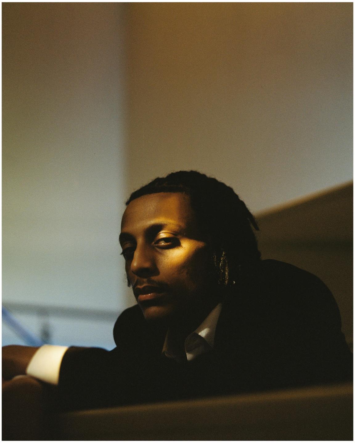 A man with dreadlocks is looking towards the viewer, with a beam of light illuminating part of his face.