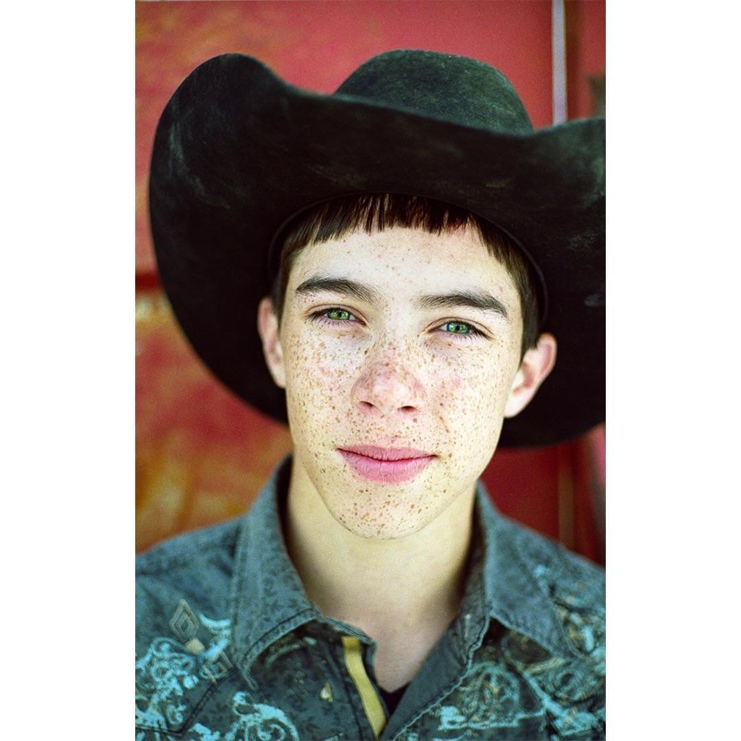 A boy with freckles and green eyes wears a cowboy hat and a patterned shirt.