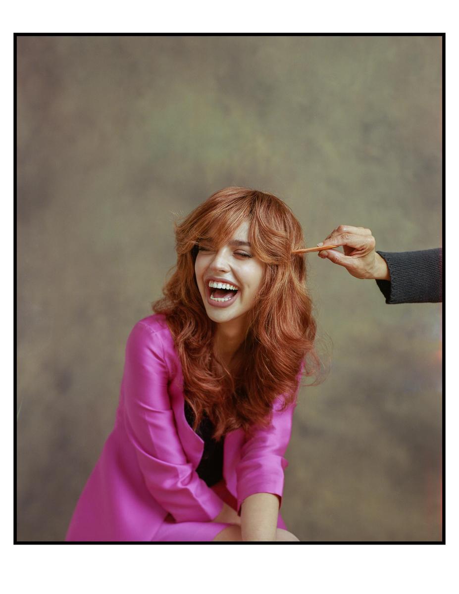 A woman with long red hair laughs while a hand holds a comb to her hair.