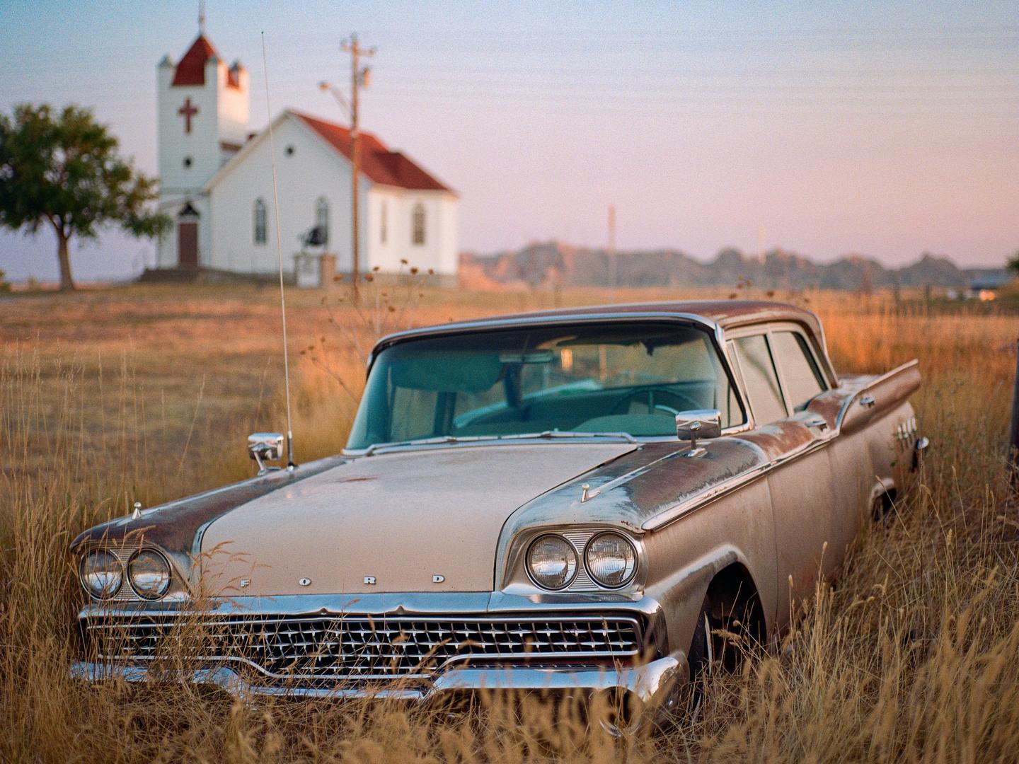 An old car sits in a field of tall grass with a white church in the background.