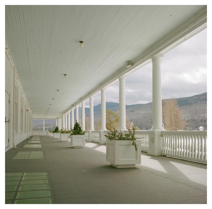 A long white porch with columns and planters stretches into the distance, overlooking a lake and mountains under a cloudy sky.