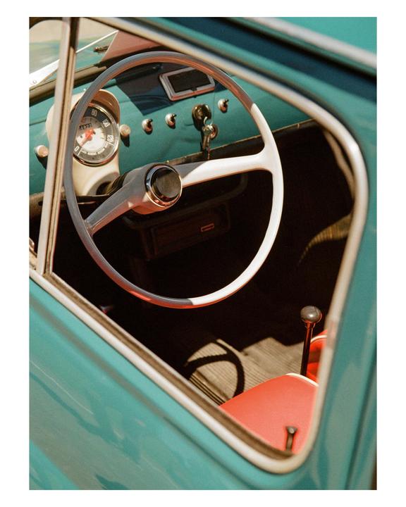 The interior of a vintage turquoise car is visible through an open window, showing the steering wheel, dashboard, and a red seat.