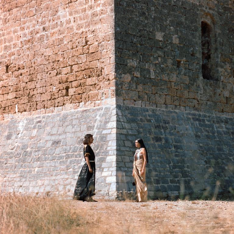 Two women in traditional clothing stand facing each other in front of a stone building.