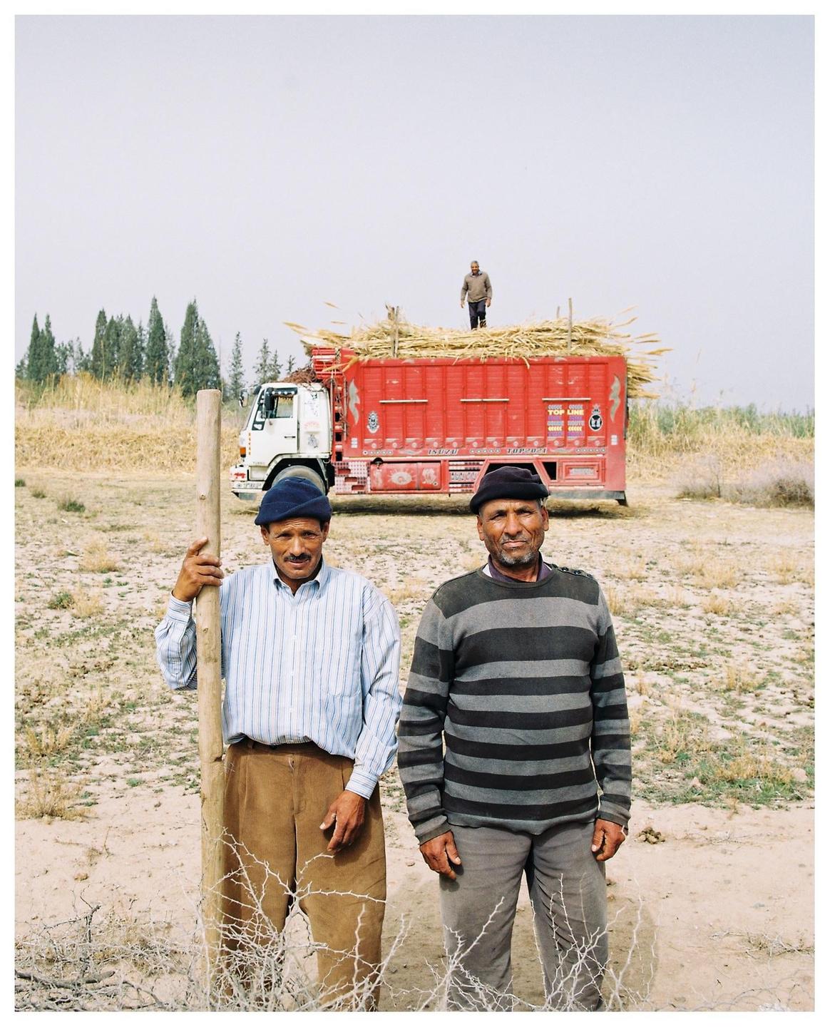 Two men stand in a field with a truck loaded with crops in the background.