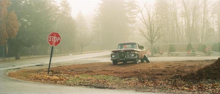 A red stop sign stands next to a road with a vintage truck parked on a dirt patch, all shrouded in fog.
