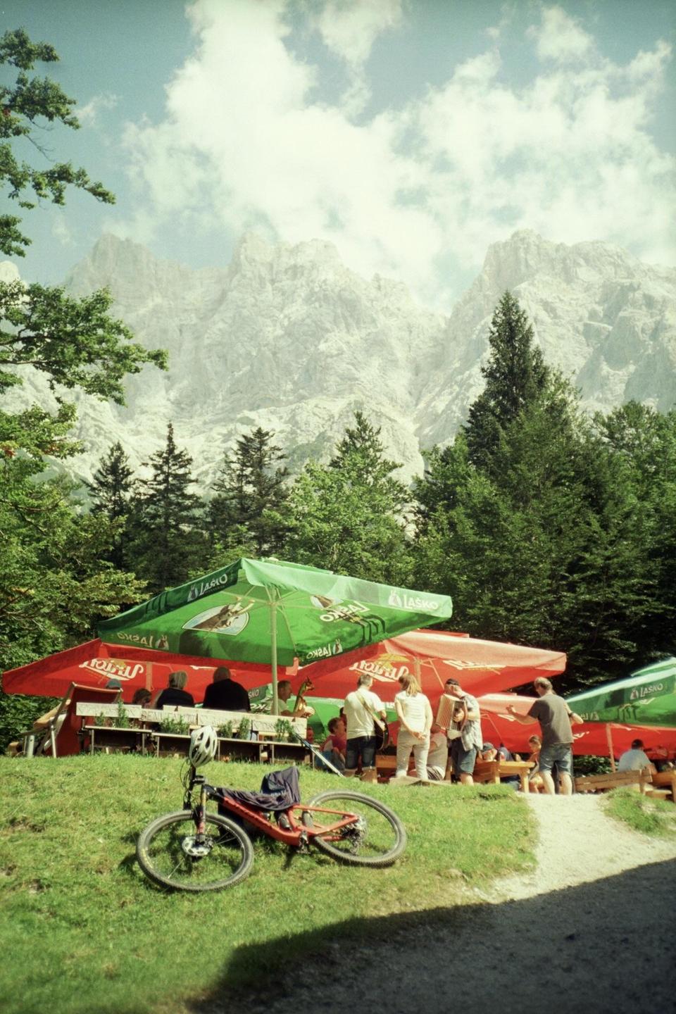 A red bicycle lies on a grassy hill in front of a restaurant with red and green umbrellas, with a large mountain and forest in the background.