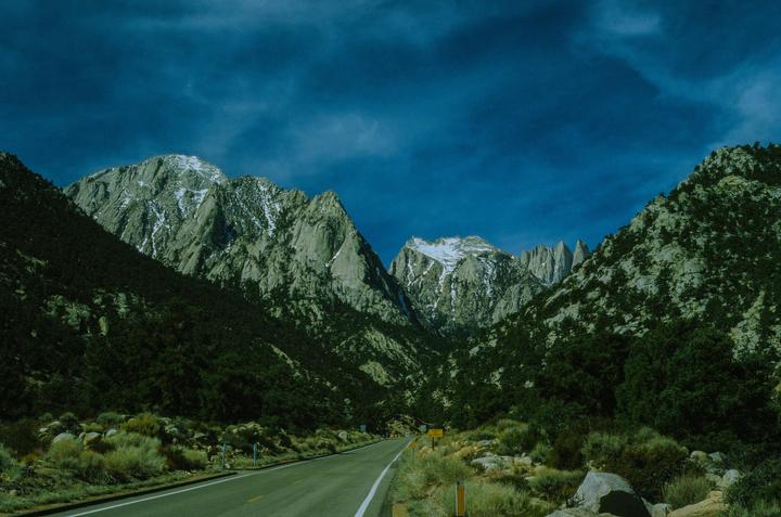 A road leads through a valley with mountains on either side, some with snow-capped peaks.