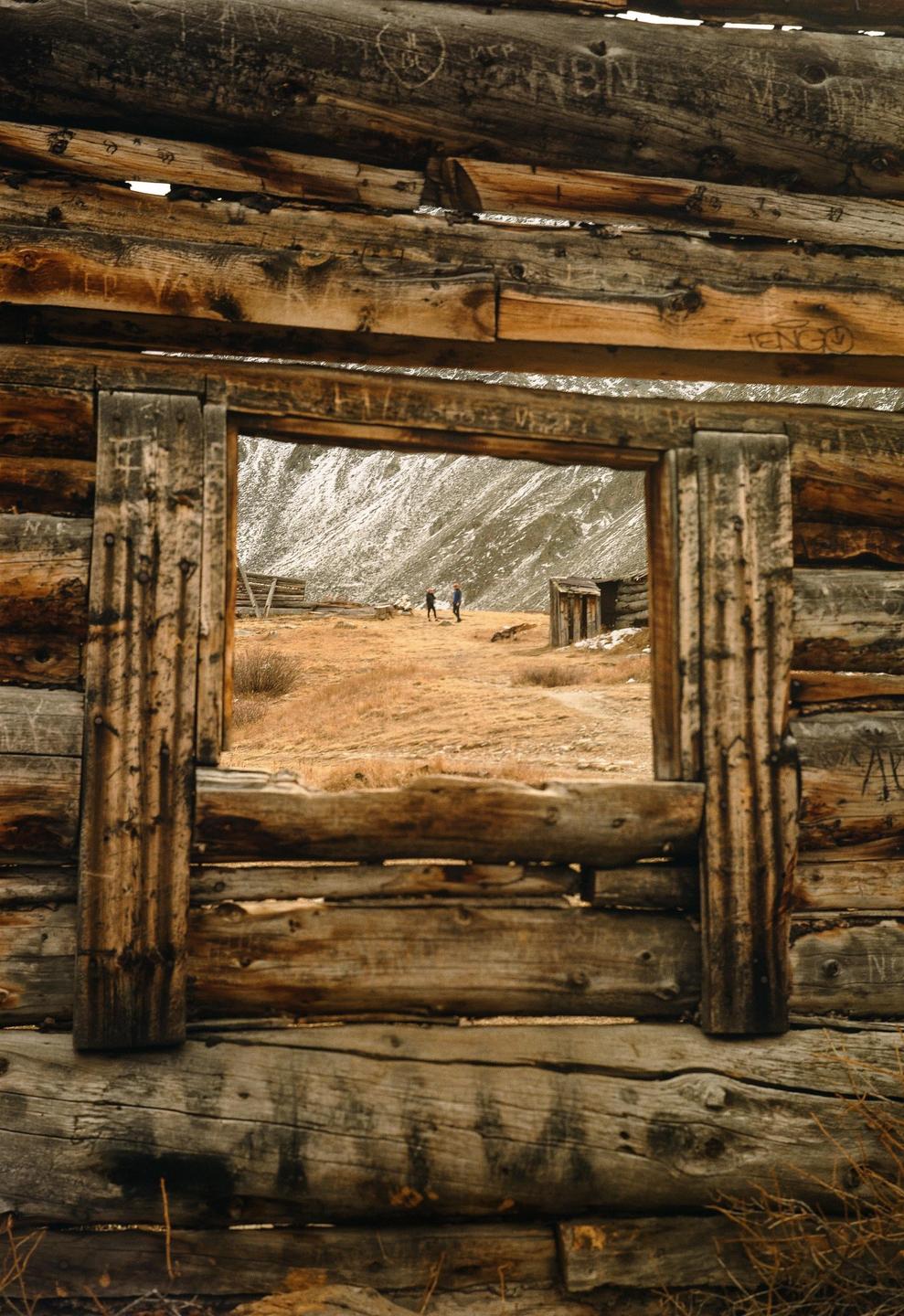 Two people walk across a field of dry grass towards a snowy mountain, viewed through a window frame of a wooden cabin.