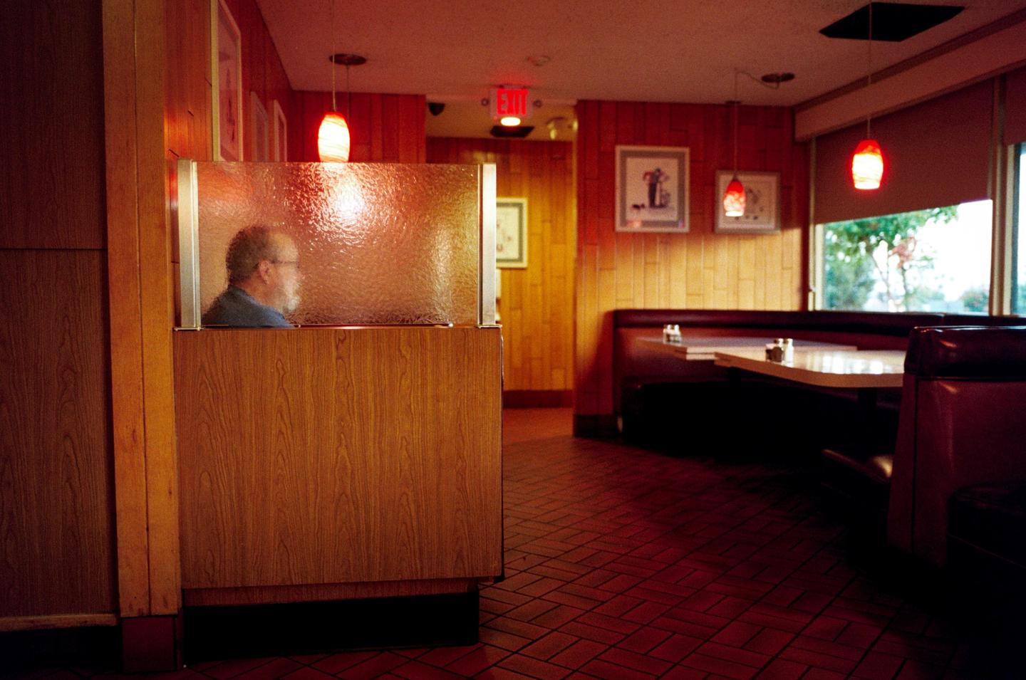 A man sits in a diner booth with red lighting and wood paneling.
