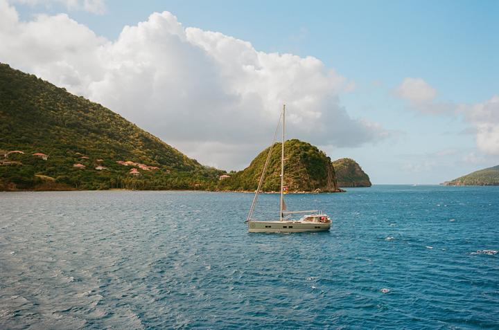 A sailboat floats on blue water with green, hilly islands in the background under a partly cloudy sky.