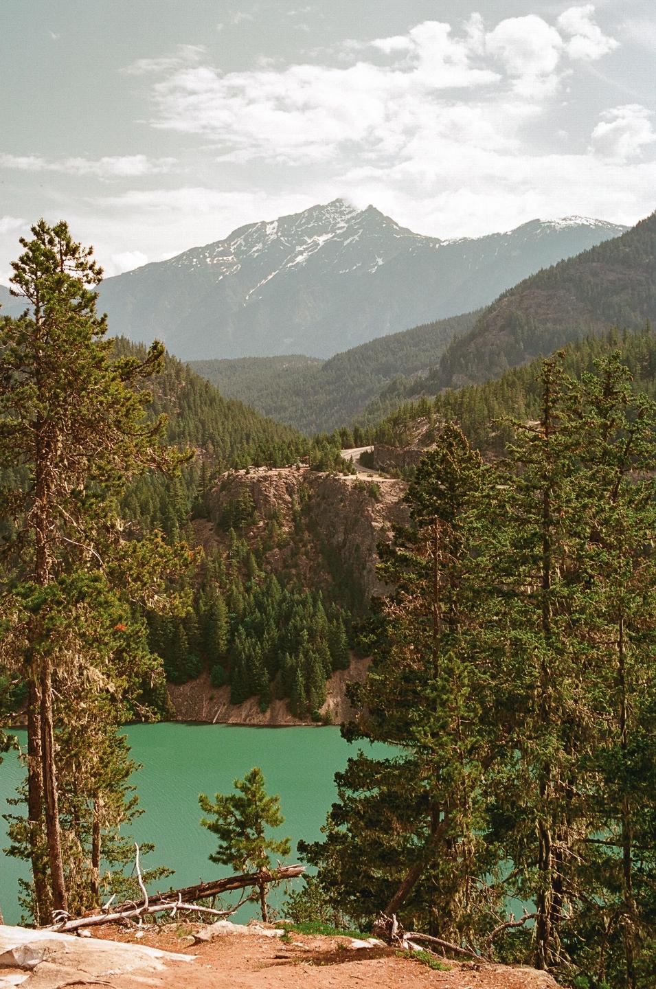 A vibrant green lake is nestled among a dense forest with snow-capped mountains in the distance under a cloudy sky.