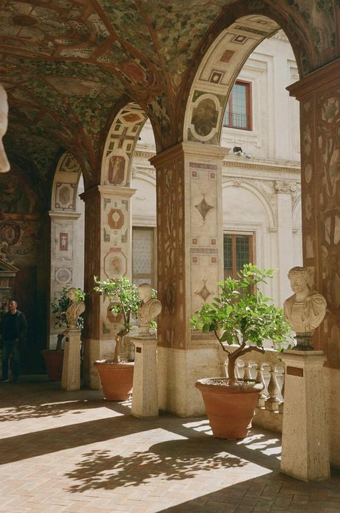A man stands in a sunlit arched hallway adorned with painted murals, potted plants, and busts.