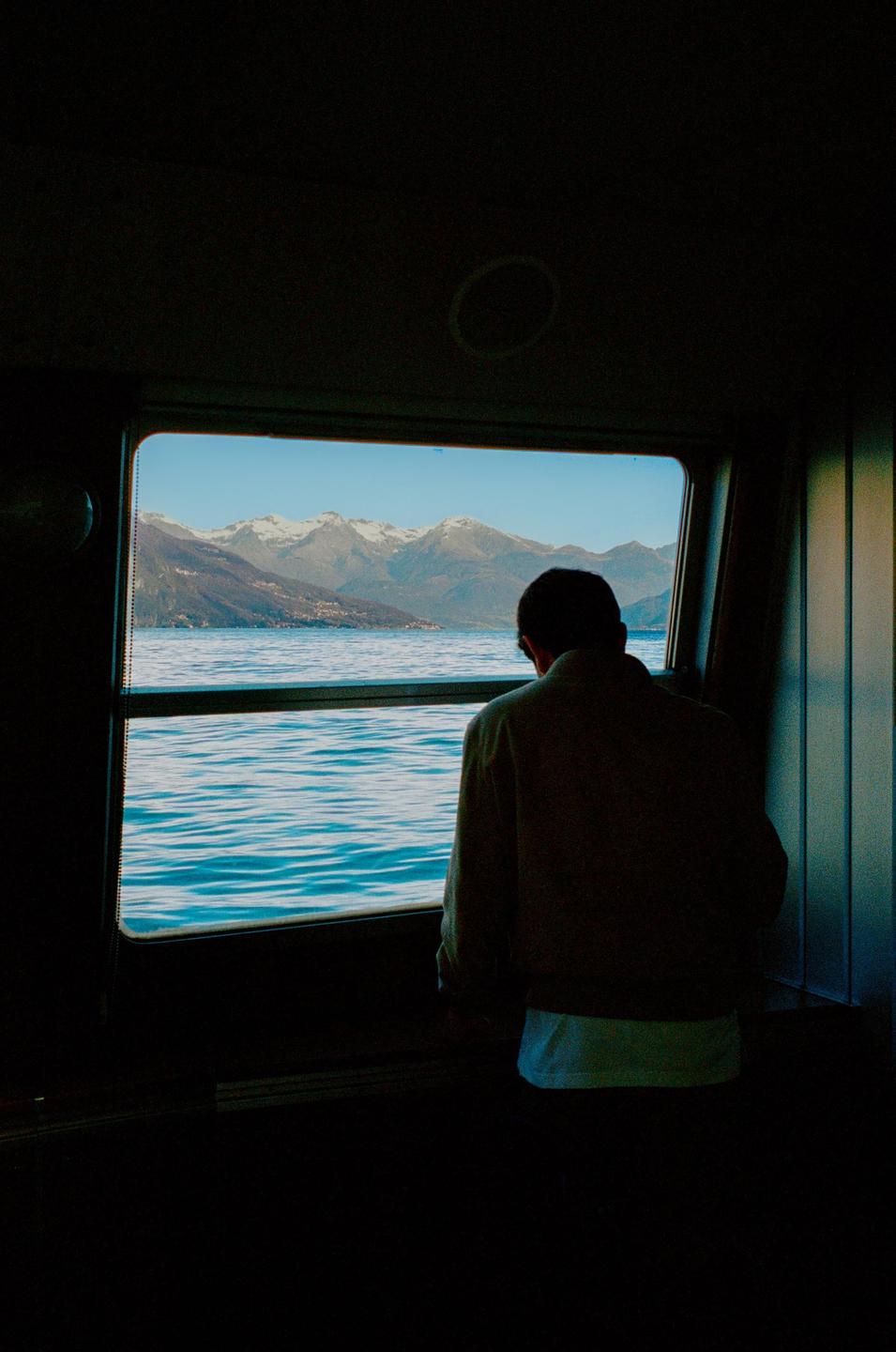 A man looks out a ferry window at a lake and snow-capped mountains.