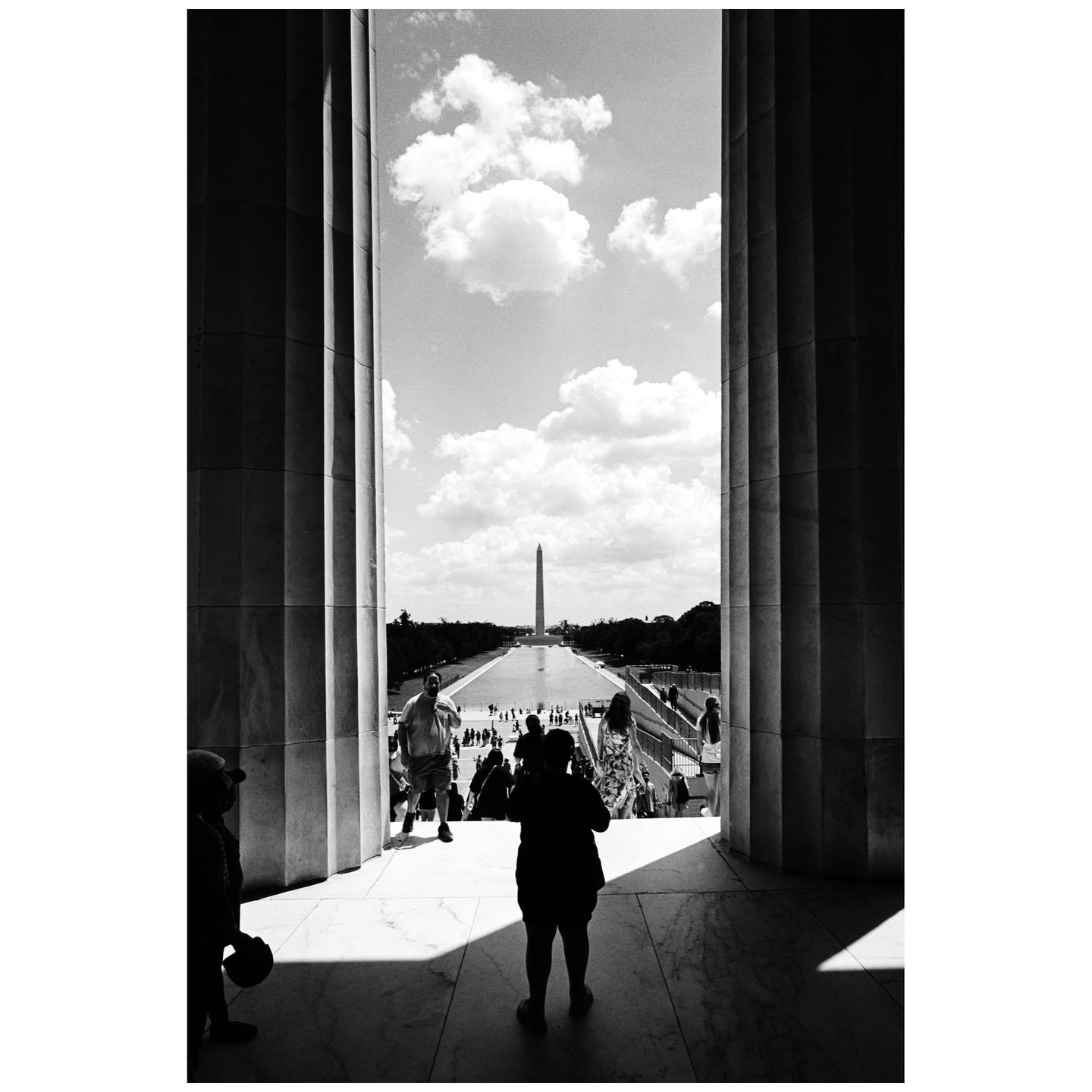 The Washington Monument is visible in the distance between two large columns, with people walking on steps in the foreground.