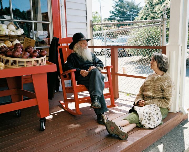 A man with a long white beard sits in a red rocking chair next to a woman sitting on a porch with baskets of onions.