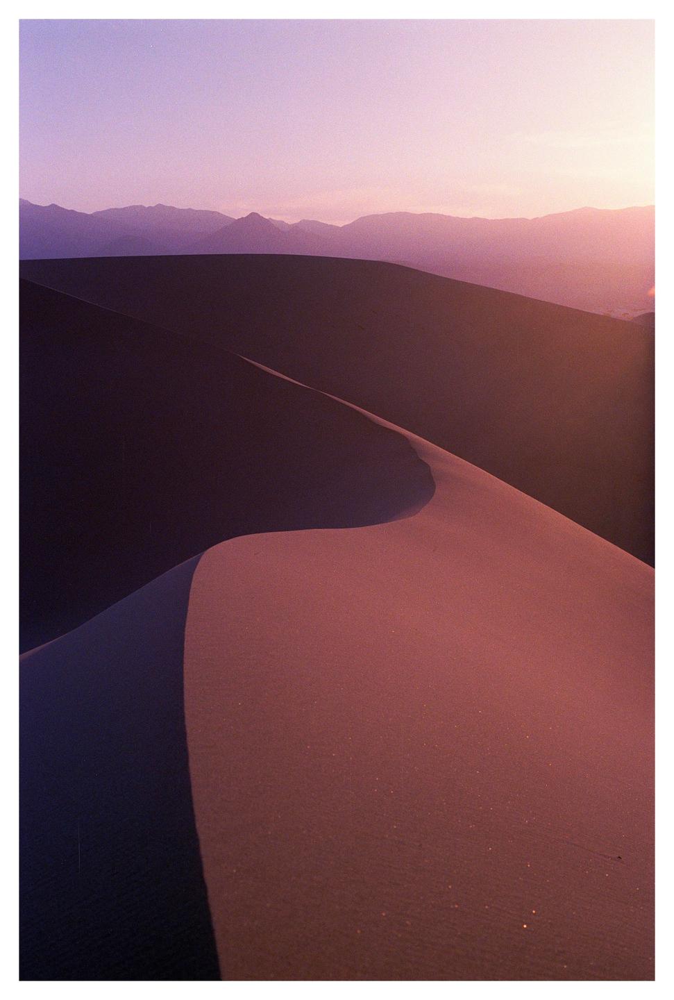 A desert landscape shows sand dunes and mountains under a purple sky at dusk.