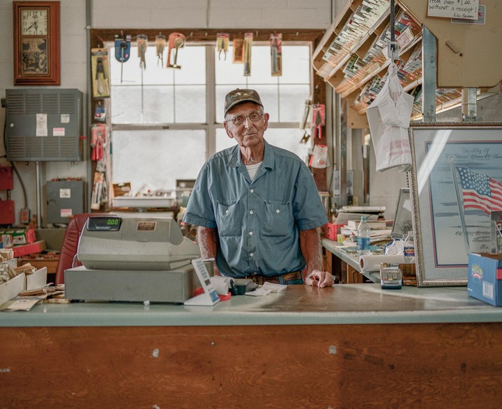 An elderly man stands behind a counter in a general store.