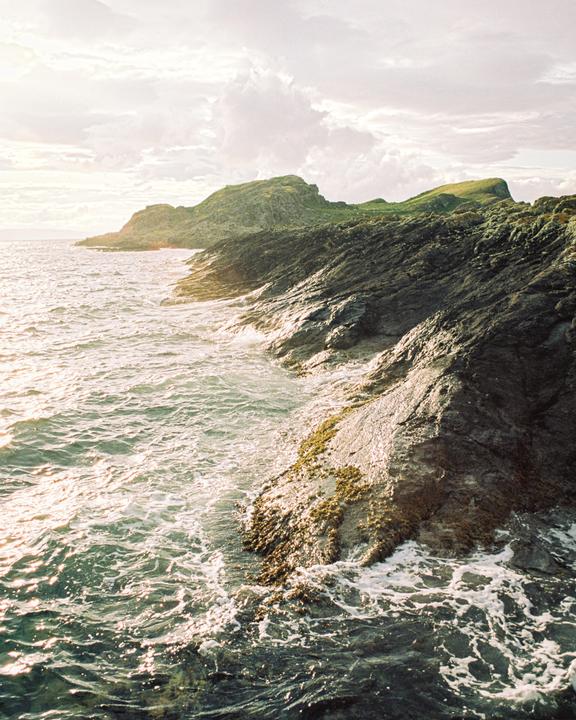 Waves crash against a rocky coastline with a green hill in the background under a cloudy sky.