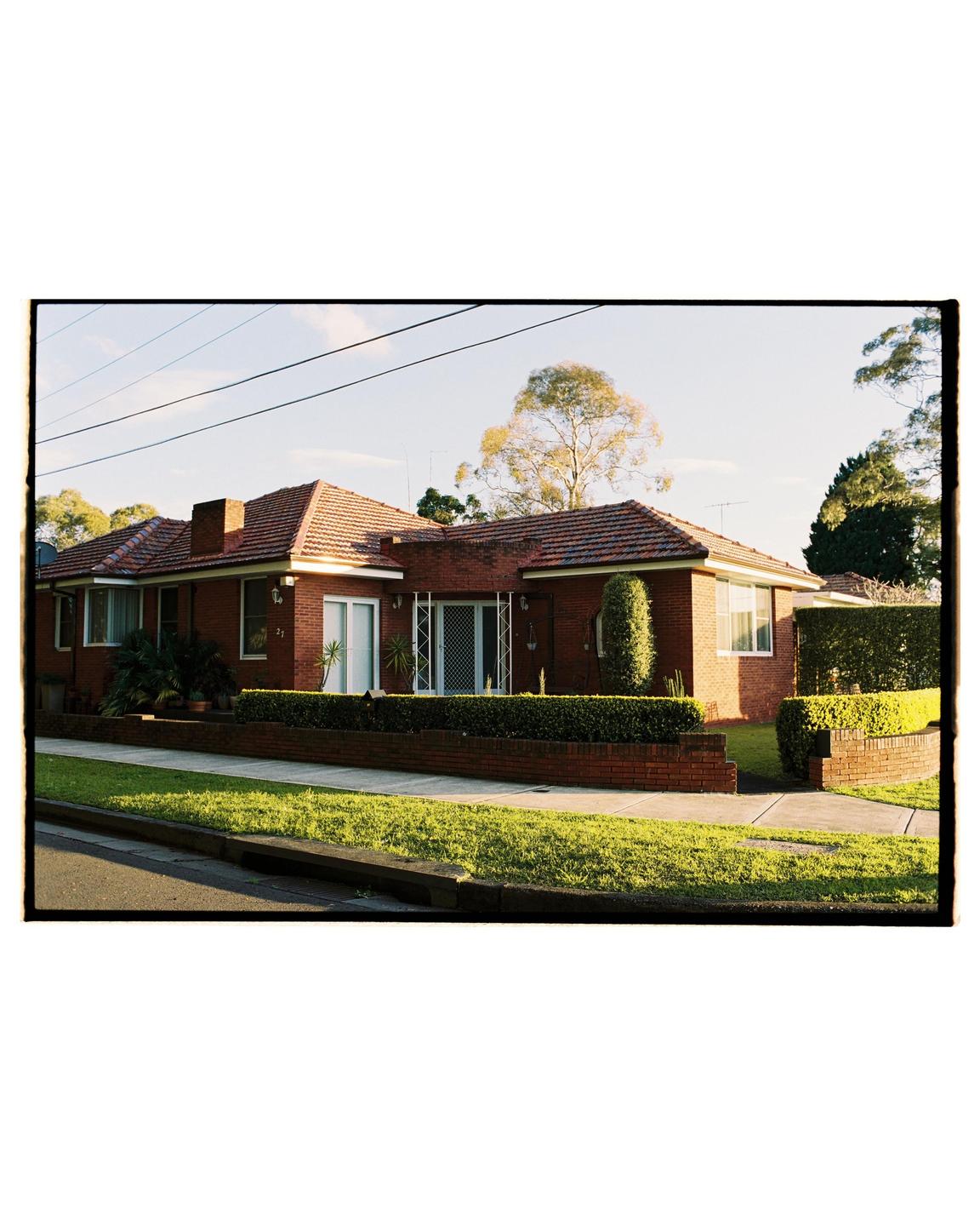 A brick house with a tiled roof is seen from the street, with green hedges and a sidewalk in front.