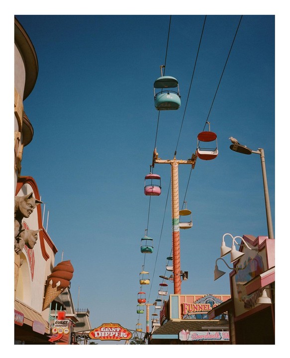 A row of colorful gondolas hang from a cable ride above an amusement park with a clear blue sky.