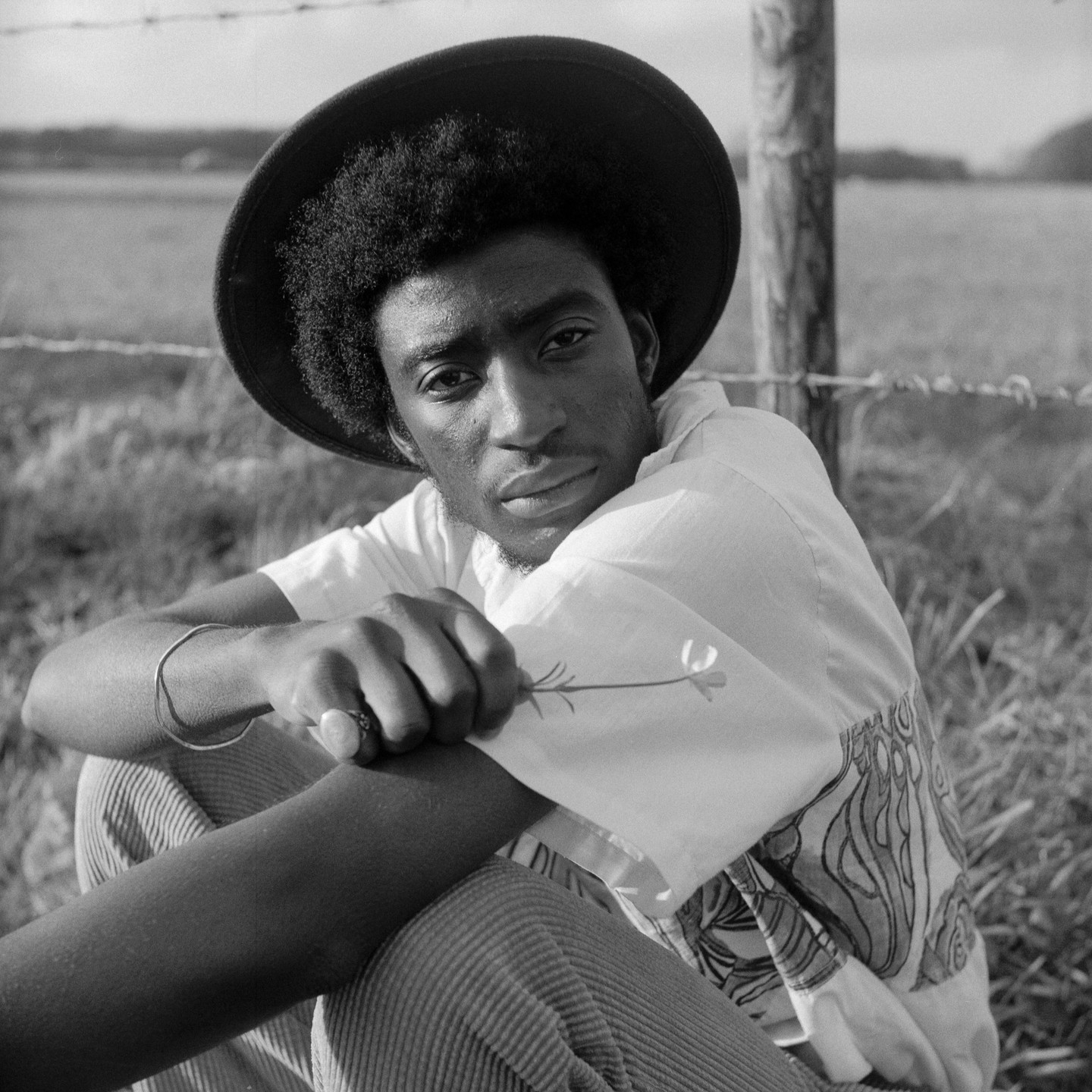 A man wearing a hat and a patterned shirt sits in a grassy field next to a barbed wire fence.