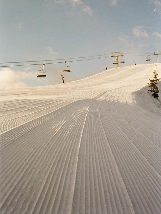 A ski slope with fresh corduroy snow is visible under a clear sky with ski lifts overhead.