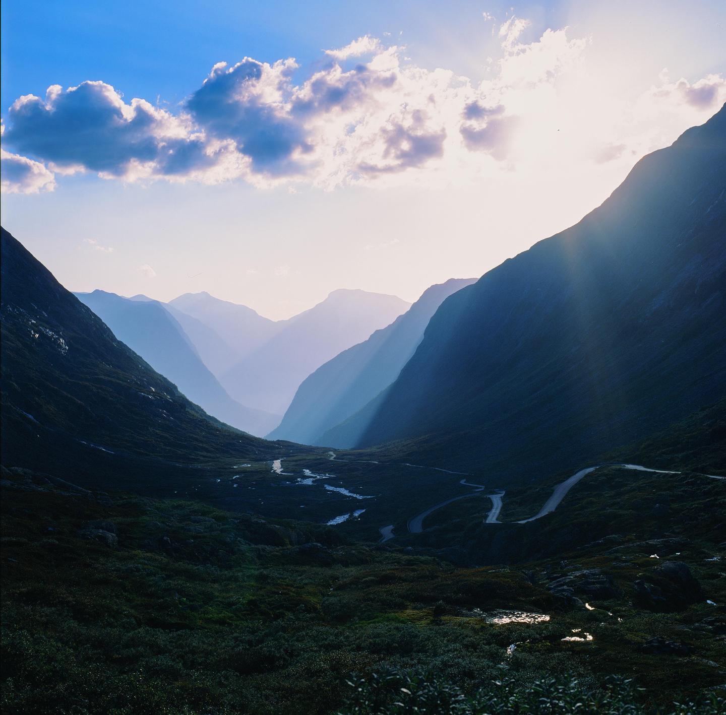 Sunlight streams over a mountain valley with a winding road and river.